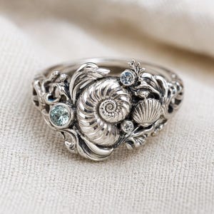 May include: A silver ring with an intricate seashell design. The ring features a large nautilus shell, a small scallop shell, and several small, round, light blue gemstones. The ring is set on a neutral background.