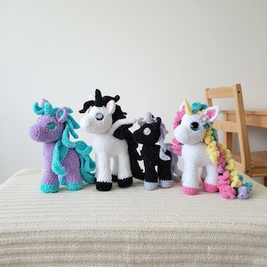 May include: Four crocheted unicorn figurines in various colors. One is purple with teal accents, another is white with black, a third is black with gray, and the last is white with a rainbow mane and pink hooves. They are standing on a white surface.