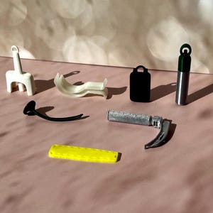 May include: Assortment of small, medical-themed objects. Includes a white, three-legged stand, a curved white piece, a black handle, a black rectangular case, a silver and green cylinder, a silver medical tool, and a yellow rectangular piece.