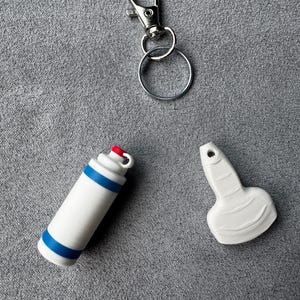May include: A silver metal keychain with a lobster clasp and a split ring, a white cylindrical bottle-shaped charm with two horizontal blue stripes and a red cap, and a white flat, spade-shaped charm with a small hole at the top for attachment.