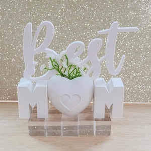 May include: A white decorative sign featuring the word Best in cursive script above the letters M and M, with a white heart-shaped vase in the center containing small green sprigs. The entire structure rests on a clear rectangular base.