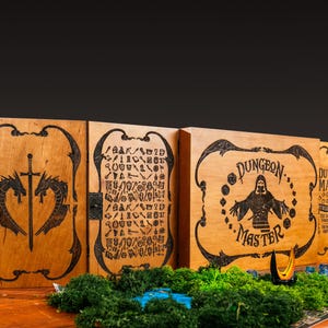 Op de afbeelding: Houten Dungeon Master-spelset met gegraveerde ontwerpen. De set bevat meerdere panelen met ingewikkelde kunstwerken, waaronder een zwaard- en drakenmotief, en de woorden "Dungeon Master". De set wordt weergegeven op een houten tafel met groen mos.