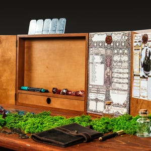 Op de afbeelding: Een houten tabletop gaming organizer met een centraal compartiment, een lade en zijpanelen. De organizer heeft een karakterblad, dobbelstenen, een pen en een leren notitieboek. De scène is versierd met groen mos en kleine glazen flesjes.