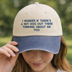May include: A beige and blue baseball cap with the text "I WONDER IF THERE'S A HOT DOG OUT THERE THINKING ABOUT ME TOO" in navy blue. The cap has a curved peak and a button on top.