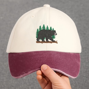 Può includere: Cappellino da baseball color crema con visiera bordeaux. Il cappellino presenta un orso nero ricamato che cammina su un tronco davanti a pini verdi. Un accessorio classico e casual.