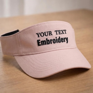 Pode incluir: Viseira rosa claro com o texto "YOUR TEXT Embroidery" bordado em preto. A viseira tem uma aba curva e uma faixa interna preta. A viseira é feita de um tecido macio e fica em uma superfície de madeira.