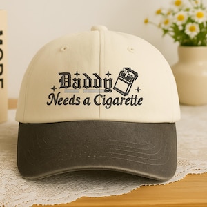 May include: A beige and dark gray baseball cap with the text "Daddy Needs a Cigarette" in black. The cap features a curved brim and a classic six-panel design. The text is accompanied by a graphic of a cigarette pack.