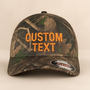 Pode incluir: Boné de beisebol camuflado com o texto bordado "CUSTOM TEXT" em laranja. O boné apresenta uma aba curva e uma etiqueta Flexfit. O padrão do boné inclui tons de verde, castanho e preto.
