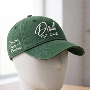 May include: A green baseball cap with the word "Dad" in white script and "EST. 2026" printed on the front. The cap also features the name "Sutton Edward Preston" embroidered on the side. The cap is displayed on a white mannequin head.
