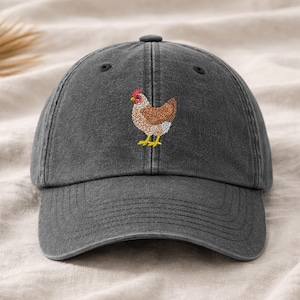 May include: A charcoal gray baseball cap featuring an embroidered chicken design. The chicken has brown, white, and red details with yellow feet. The cap has a curved brim and a button on top.