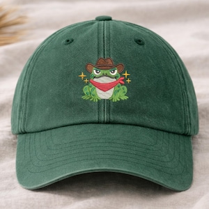 May include: A dark green baseball cap featuring an embroidered frog wearing a cowboy hat and red bandana. The frog has a stern expression and is surrounded by gold star accents. The cap has a curved brim and a fabric strap closure.