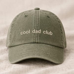 Puede incluir: Gorra de béisbol verde salvia con la inscripción bordada "cool dad club" en blanco. La gorra tiene una visera curva y un botón en la parte superior. La tela parece ser algodón lavado o similar.