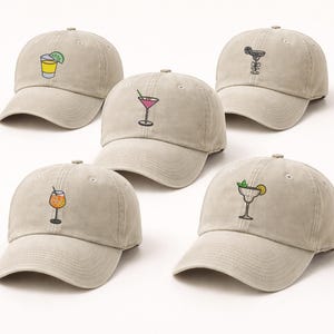 Puede incluir: Cinco gorras de béisbol beige con diseños de cócteles bordados. Cada gorra presenta un cóctel diferente: una margarita, un martini, un cóctel de naranja, un cóctel con lima y un cóctel con menta y limón.