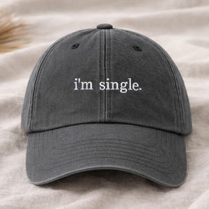 Könnte beinhalten: Anthrazitfarbene Baseballkappe mit dem gestickten Text "I'm single." Die Kappe hat einen gebogenen Schirm und einen verstellbaren Riemen auf der Rückseite. Der Hut ist aus einem weichen, strukturierten Stoff gefertigt.