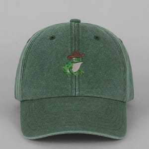 May include: Green baseball cap featuring an embroidered frog wearing a cowboy hat. The cap is a solid, muted green colour with a curved peak and adjustable strap. The frog is green and white with a brown hat.