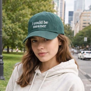May include: A dark green baseball cap with the text "i could be meaner" in white. The cap is worn by a person wearing a light grey hoodie. The background includes trees, a street, and buildings.