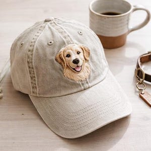 Puede incluir: Gorra de béisbol gris claro con una imagen bordada de un perro Golden Retriever. La gorra está junto a una taza de cerámica con café y un collar de perro de cuero marrón con una etiqueta.