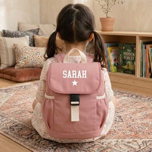 May include: A dusty pink backpack with the name "SARAH" in white and a white star. The backpack has a top handle, a black buckle, and a cream strap. The child is sitting on a patterned rug, with books in the background.