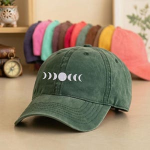 May include: A dark green baseball cap with a curved peak. The cap has a white embroidered moon phase design on the front. Other baseball caps in various colours are visible in the background.
