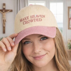 Könnte beinhalten: Beige und rosa Baseballkappe mit dem gestickten Text "MAKE HEAVEN CROWDED". Die Kappe hat einen rosa Schirm und ein gebogenes Visier. Die Mütze wird von einer Person gehalten.