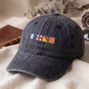 Op de afbeelding: Een donkergrijze baseballcap met geborduurde nautische vlaggen. De vlaggen zijn wit, blauw, rood, geel en oranje. De cap is gemaakt van een zacht, versleten materiaal.