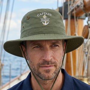 May include: An olive green bucket hat with the embroidered text "CAPTAIN Ember" and an anchor design. The hat has a wide brim, eyelets, and an adjustable chin strap. The person in the image is wearing a navy blue polo shirt.