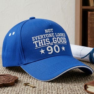 May include: A royal blue baseball cap with the text "NOT EVERYONE LOOKS THIS GOOD at 90" in white. The cap has a white trim and two white dots. A celebratory gift for a 90th birthday.