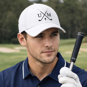Puede incluir: Gorra de béisbol blanca con las letras "UXM" y palos de golf cruzados impresos en negro. La gorra tiene una visera curva y está hecha de un material suave. Accesorio de golf.