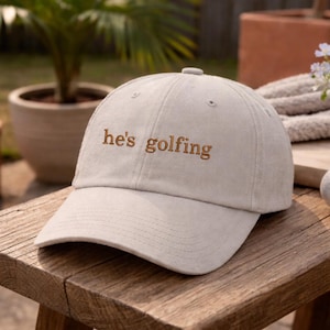 May include: A beige baseball cap with the embroidered text "he's golfing" in brown. The hat is on a wooden bench with a golf ball, a small vase of flowers, and a book. The background includes potted plants.
