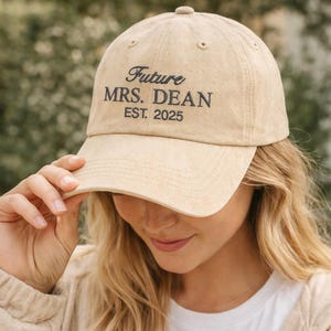 Puede incluir: Gorra de béisbol beige con la inscripción bordada "Future MRS. DEAN EST. 2025". La gorra tiene una visera curva y un botón en la parte superior. El sombrero está hecho de una tela suave y texturizada.