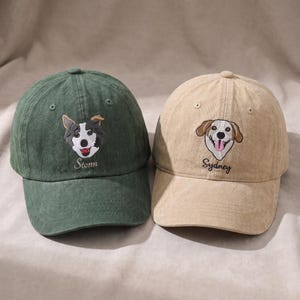 Könnte beinhalten: Zwei Baseballkappen, eine dunkelgrün und eine beige, jeweils mit einem gestickten Hundegesicht auf der Vorderseite. Die grüne Kappe zeigt einen schwarz-weißen Hund mit dem darunter gestickten Namen Storm, während die beige Kappe einen braun-weißen Hund mit dem darunter gestickten Namen Sydney zeigt.