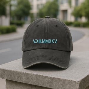 May include: A charcoal gray baseball cap with a curved brim. The cap features the Roman numeral text "V.XIII.MMXXV" in a light blue color on the front. The cap is sitting on a stone surface.