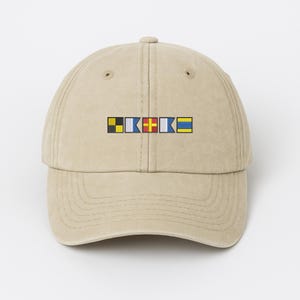 May include: A tan baseball cap featuring a series of colorful nautical signal flags embroidered across the front. The flags are in yellow, black, blue, red, and white. The cap has a curved brim and a classic six-panel design.