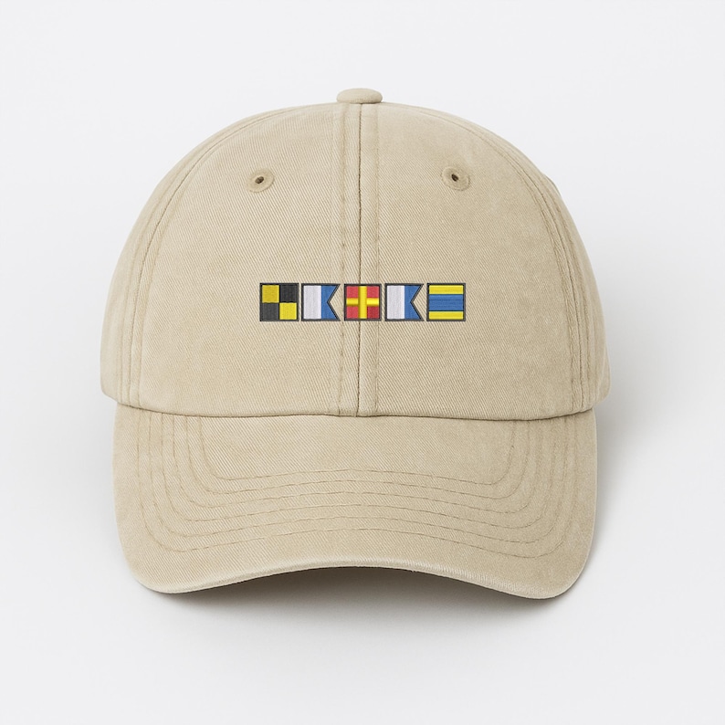 May include: A tan baseball cap featuring a series of colorful nautical signal flags embroidered across the front. The flags are in yellow, black, blue, red, and white. The cap has a curved brim and a classic six-panel design.