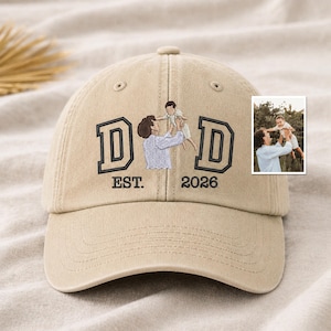 May include: A beige baseball cap embroidered with the word "DAD" in black. The cap features an embroidered image of a person holding a child, with the text "EST. 2026" below. A small photo is visible on the right.
