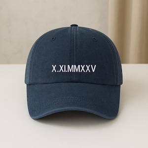 May include: A navy blue baseball cap with the Roman numeral date X.XI.MMXXV printed in white. The cap has a curved brim and a button on top. The hat is made of a soft, durable material.