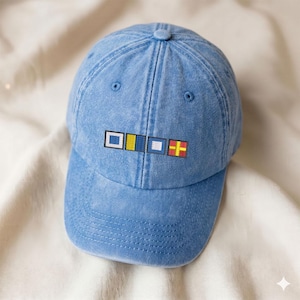 May include: A light blue baseball cap with a curved brim. The cap features embroidered nautical signal flags in white, yellow, blue, and red. The hat is made of a soft, washed denim material.