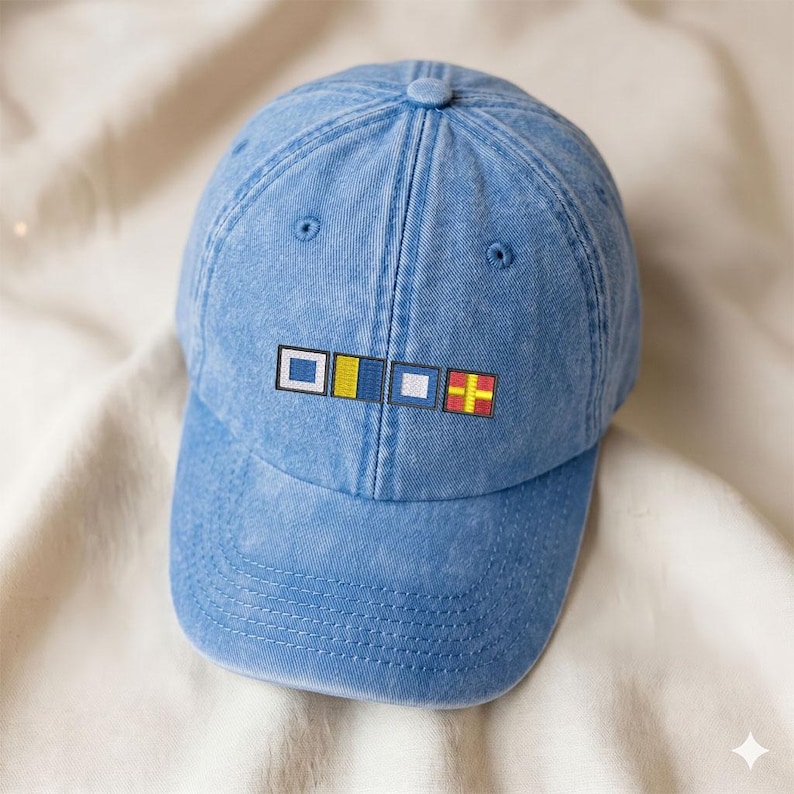 May include: A light blue baseball cap with a curved brim. The cap features embroidered nautical signal flags in white, yellow, blue, and red. The hat is made of a soft, washed denim material.