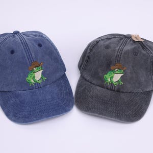 May include: Two baseball caps, one navy blue and one charcoal grey, are shown. Each cap has an embroidered green frog wearing a brown cowboy hat. The caps have a distressed, vintage appearance.