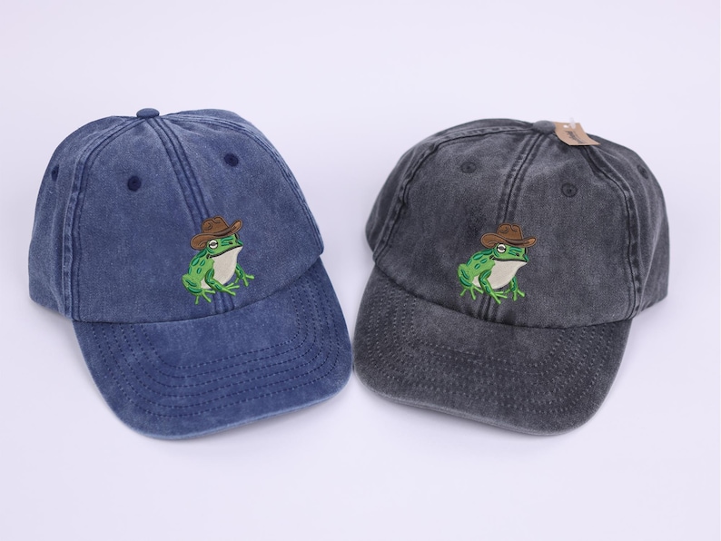 May include: Two baseball caps, one navy blue and one charcoal grey, are shown. Each cap has an embroidered green frog wearing a brown cowboy hat. The caps have a distressed, vintage appearance.