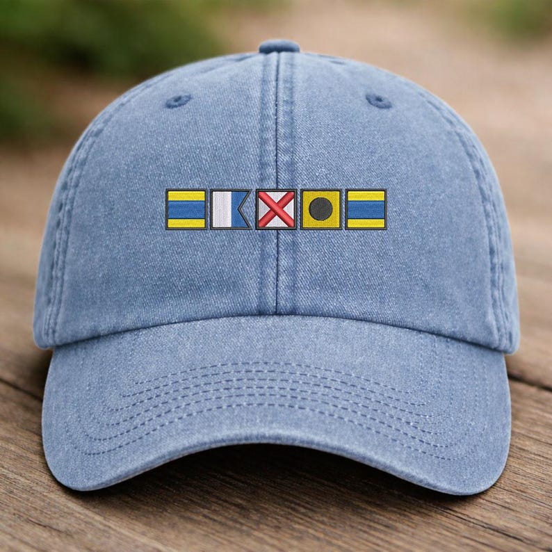 May include: A light blue baseball cap with a curved brim. The cap features an embroidered design of nautical signal flags in yellow, white, red, and black. The cap is made of a soft, textured fabric.