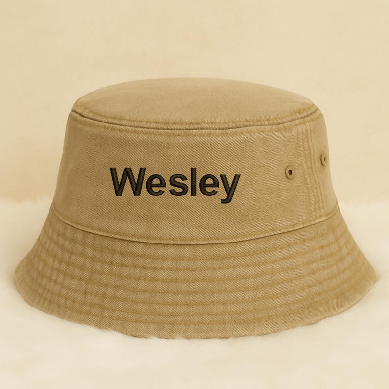May include: Tan bucket hat with the name "Wesley" embroidered in black. The hat has a classic design with a slightly sloped brim and a structured crown. The hat is made of a durable fabric and features a simple, casual style.