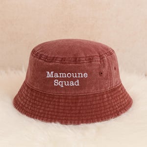 May include: A maroon bucket hat with the text "Mamoune Squad" embroidered in white. The hat is made of a soft, textured fabric and has a classic bucket hat design.