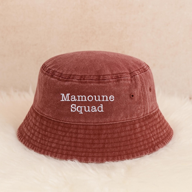 May include: A maroon bucket hat with the text "Mamoune Squad" embroidered in white. The hat is made of a soft, textured fabric and has a classic bucket hat design.