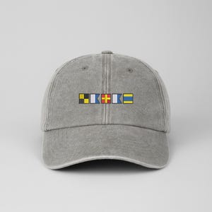 May include: A gray baseball cap with a curved brim. The cap features a design of colorful nautical signal flags embroidered across the front. The flags are yellow, black, blue, red, and white.