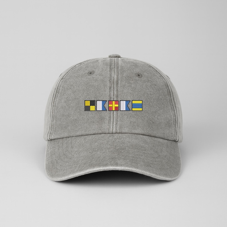 May include: A gray baseball cap with a curved brim. The cap features a design of colorful nautical signal flags embroidered across the front. The flags are yellow, black, blue, red, and white.