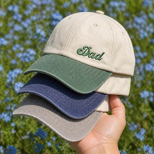 Pode incluir: Uma pilha de quatro bonés de beisebol em bege, verde, azul marinho e cinza. O boné superior tem a palavra "Dad" bordada em verde. Os bonés são segurados em uma mão contra um fundo de flores azuis.