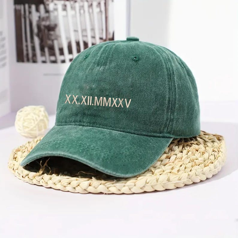 May include: A green baseball cap with the Roman numeral date XX.XII.MMXXV embroidered in white. The cap is made of a soft material and sits on a woven straw mat.