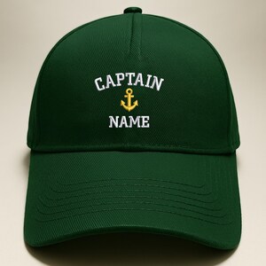 May include: A dark green baseball cap with the word "CAPTAIN" embroidered in white above a gold anchor and the word "NAME" in white. The cap has a curved brim and a rounded crown.