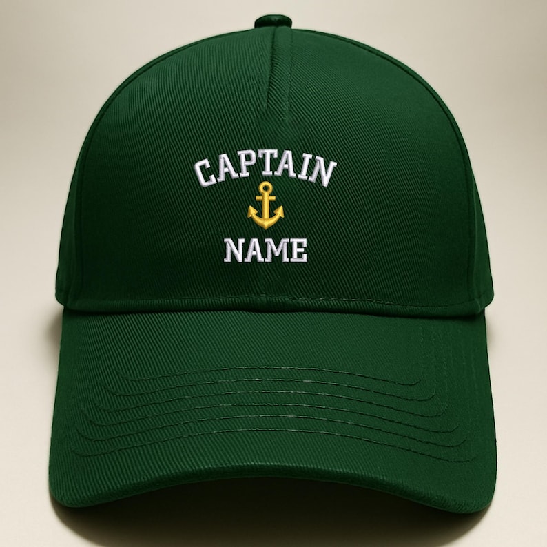 May include: A dark green baseball cap with the word "CAPTAIN" embroidered in white above a gold anchor and the word "NAME" in white. The cap has a curved brim and a rounded crown.